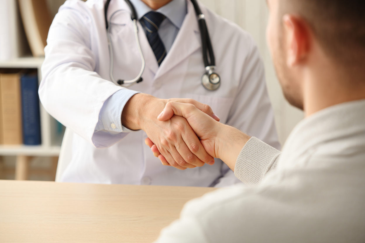 Врач в белом халате со стетоскопом пожимает руку пациенту за столом — начало принудительного лечения алкоголизма